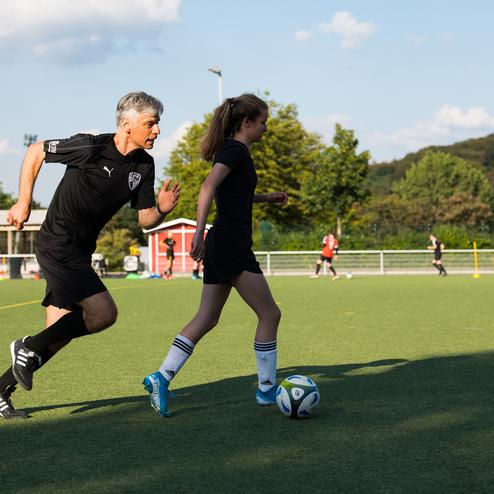 Trainer und Spielerin laufen auf einem Fu&szlig;ballfeld im Training, ein Ball liegt nahe am Boden.