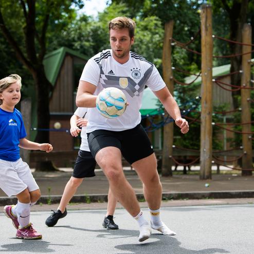 Ein Junge in einem blauen Trikot spielt Fu&szlig;ball mit einem jungen Mann in einem wei&szlig;en Trikot auf einem Sportplatz.