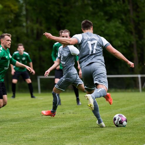 Ein Fu&szlig;ballspieler in grauem Trikot schie&szlig;t den Ball, w&auml;hrend zwei Gegner ihn stoppen wollen. Hintergrund mit B&auml;umen.