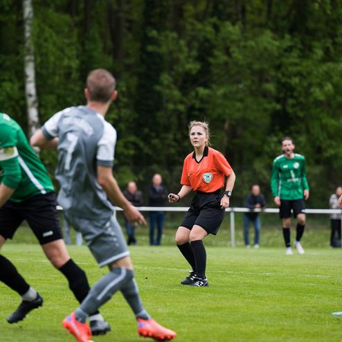 Spielszene auf einem Fu&szlig;ballplatz, Schiedsrichterin in orange mit Pfeife, Spieler in grauen und gr&uuml;nen Trikots.