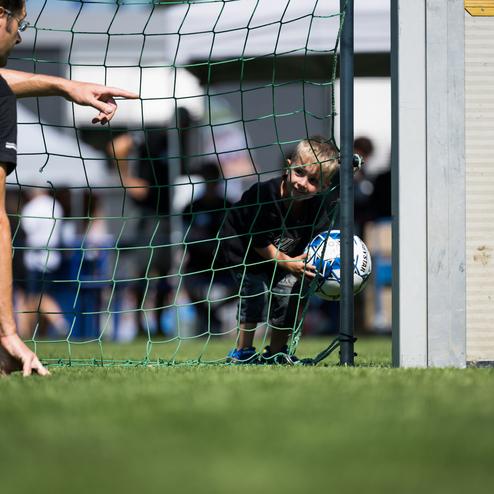 Ein Kind schaut durch das Tor und h&auml;lt einen Fu&szlig;ball, w&auml;hrend ein Erwachsener auf das Tor zeigt.