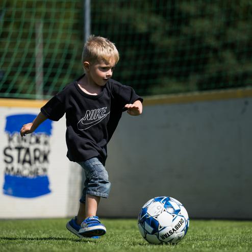 Junge in schwarzem T-Shirt kickt einen Fu&szlig;ball auf einer gr&uuml;nen Wiese, im Hintergrund eine Spielfeldbegrenzung.