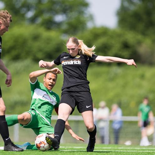 Drei Fu&szlig;ballspieler auf einem Rasenplatz, w&auml;hrend ein Spieler im gr&uuml;nen Trikot auf dem Boden liegt und der andere springt.