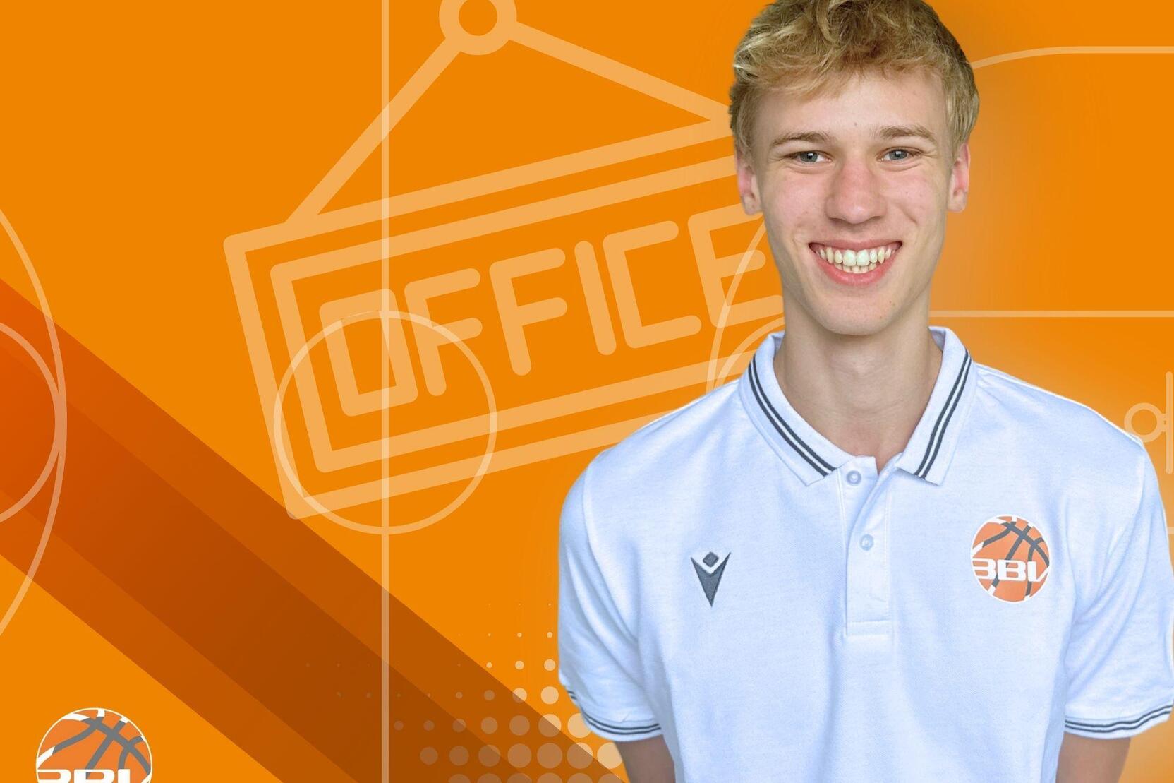 Junger Mann mit kurzem, blondem Haar, l&auml;chelt vor orangefarbenem Hintergrund mit &bdquo;OFFICE&ldquo;-Schild und Basketball-Logo.