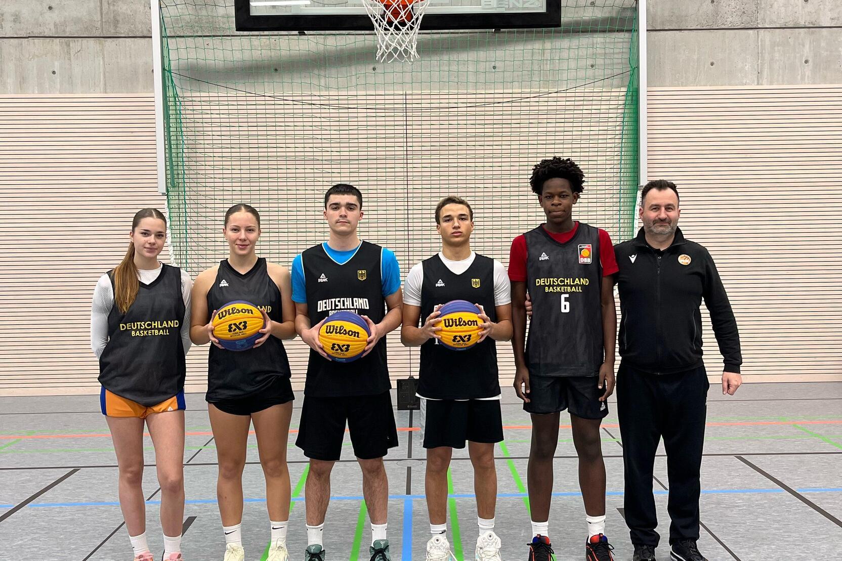 Sechs Basketballspieler in Sportkleidung stehen vor einem Basketballkorb in einer Halle und halten B&auml;lle in den H&auml;nden.