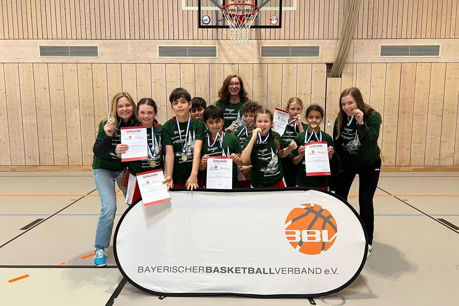 Gruppenfoto von Jugendlichen mit Medaillen und Urkunden vor einem Basketballkorb, gemeinsam mit einer Trainerin.