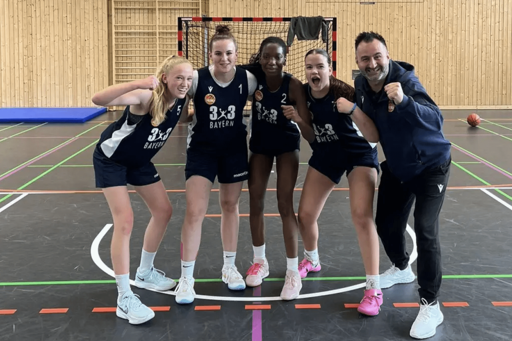 F&uuml;nf Basketballspielerinnen und ihr Trainer stehen jubelnd in Sportkleidung in einer Halle hinter einem Basketballkorb.