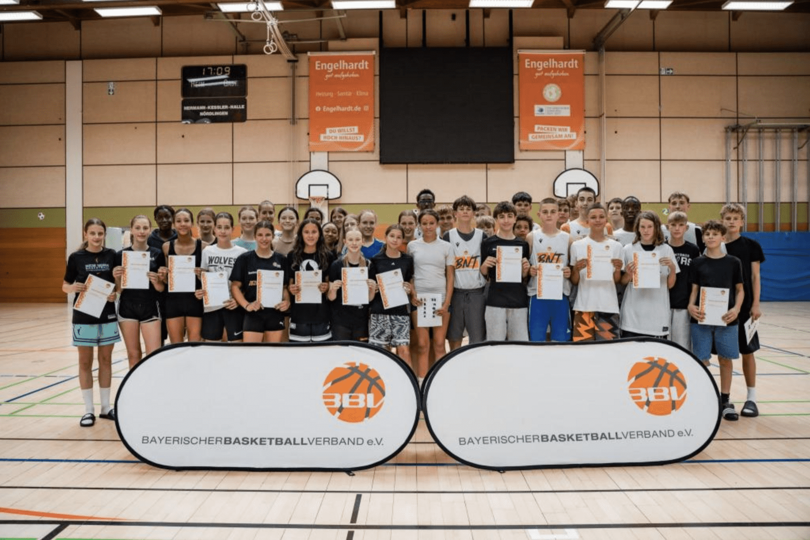 Gruppe von Jugendlichen mit Urkunden in einer Sporthalle, vor Bannern des Bayerischen Basketballverbands.