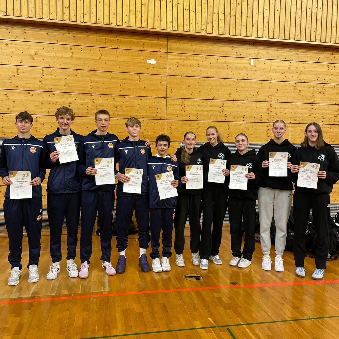 Gruppe von zehn Jugendlichen in Sportkleidung h&auml;lt Urkunden in einer Turnhalle mit Holzverkleidung.