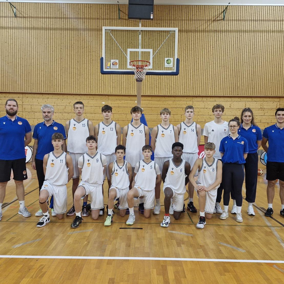 Mannschaftsfoto einer Jugend-Basketballmannschaft mit Trainern in Sportkleidung auf einem Basketballplatz.