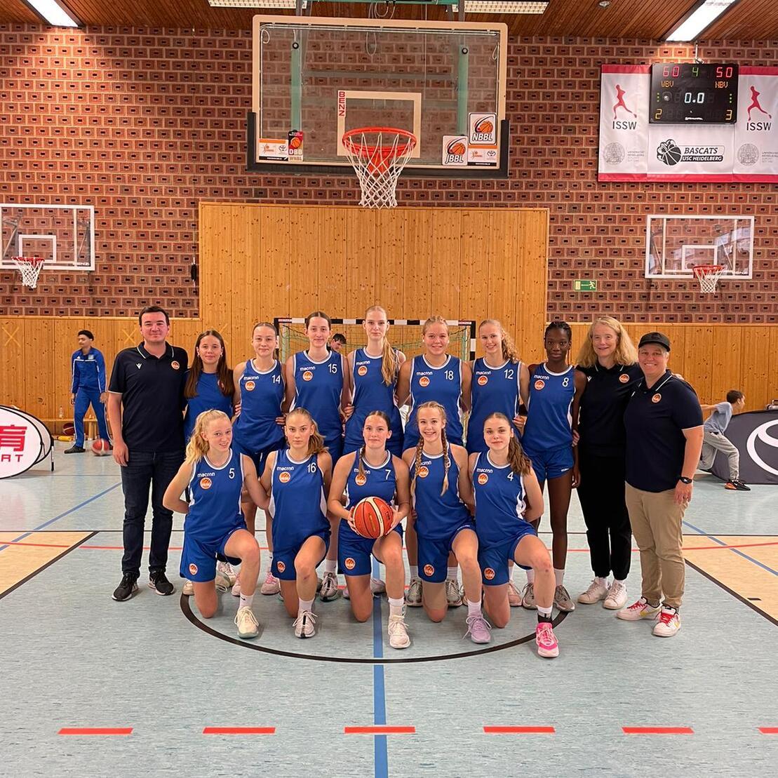 Foto einer Basketballmannschaft in blauen Trikots, stehend und kniend, mit Trainer in einer Sporthalle.