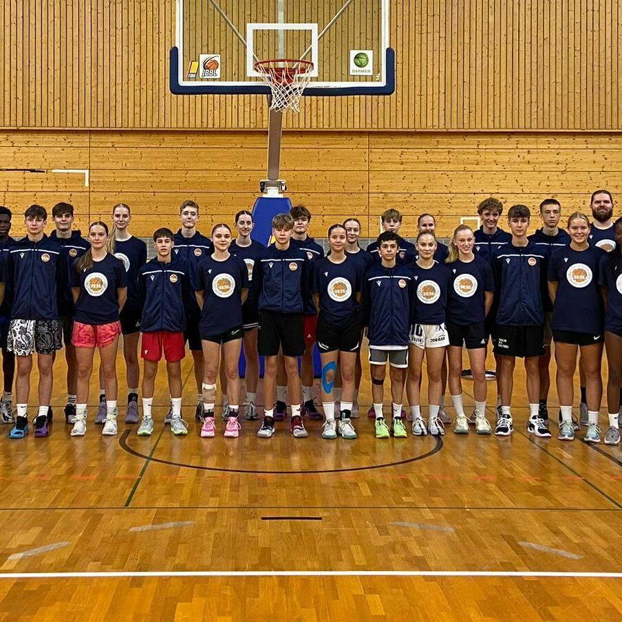Gruppe von Basketballspielern und -trainern in Sportbekleidung, aufgestellt in einer Sporthalle vor einem Basketballkorb.