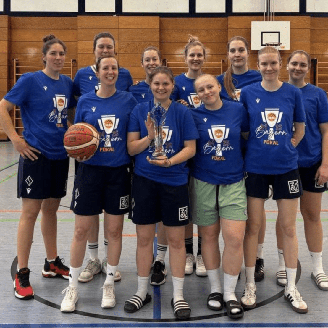 Zehn Basketballspielerinnen posieren in blauen T-Shirts, eine h&auml;lt den Pokal in der Mitte. Sporthalle im Hintergrund.