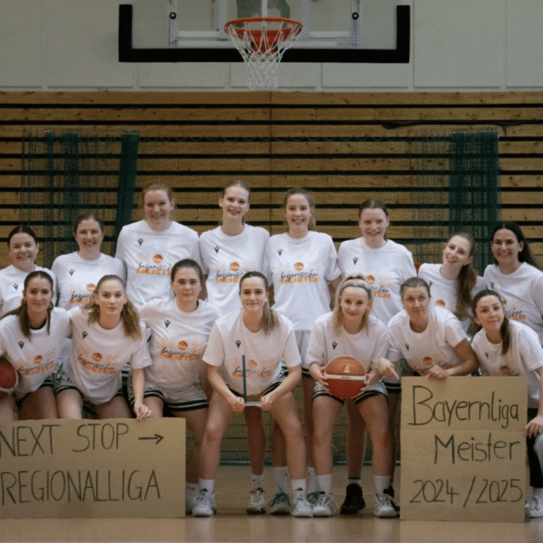 Frauen-Basketballmannschaft posiert in T-Shirts vor einem Basketballkorb, mit Schildern zu Regional- und Bayernliga-Meisterschaft.