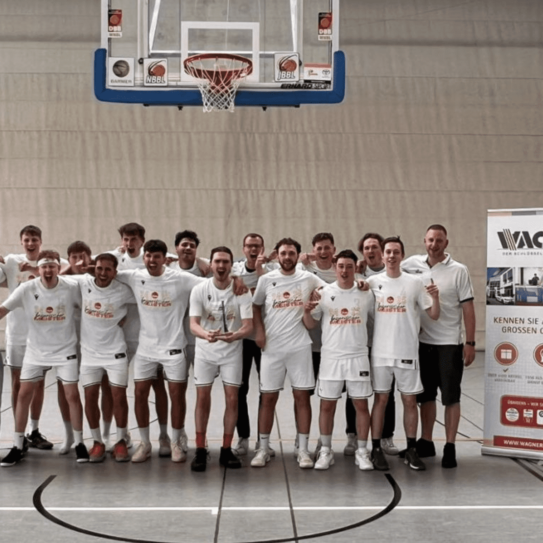 Gruppe von Basketballspielern in wei&szlig;en Trikots posiert vor einem Werbebanner in einer Sporthalle.