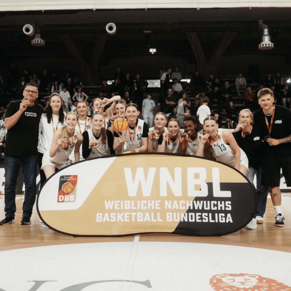 Feiernde Basketballmannschaft mit Medaillen und Pokal vor einem Banner der WNBL in einer Halle.