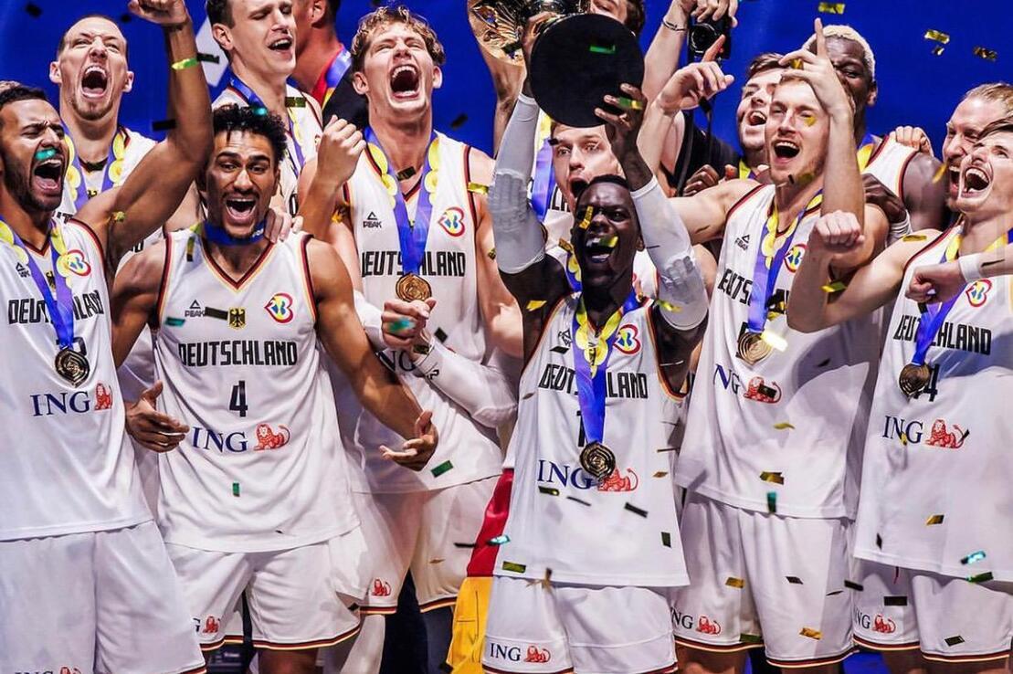 Feiernde deutsche Basketballmannschaft hebt den Pokal, w&auml;hrend Konfetti herabf&auml;llt und Medaillen um den Hals h&auml;ngen.