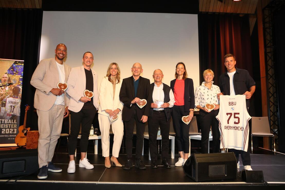Gruppenschnappschuss von acht Personen auf einer B&uuml;hne, jede h&auml;lt ein herzf&ouml;rmiges Lebkuchenherz und l&auml;chelt.