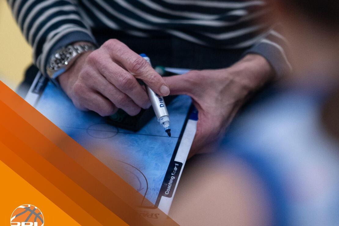 Ein Coach hält einen Marker über einem Basketballspiel-Plan auf einem Tisch, um Taktiken zu skizzieren.