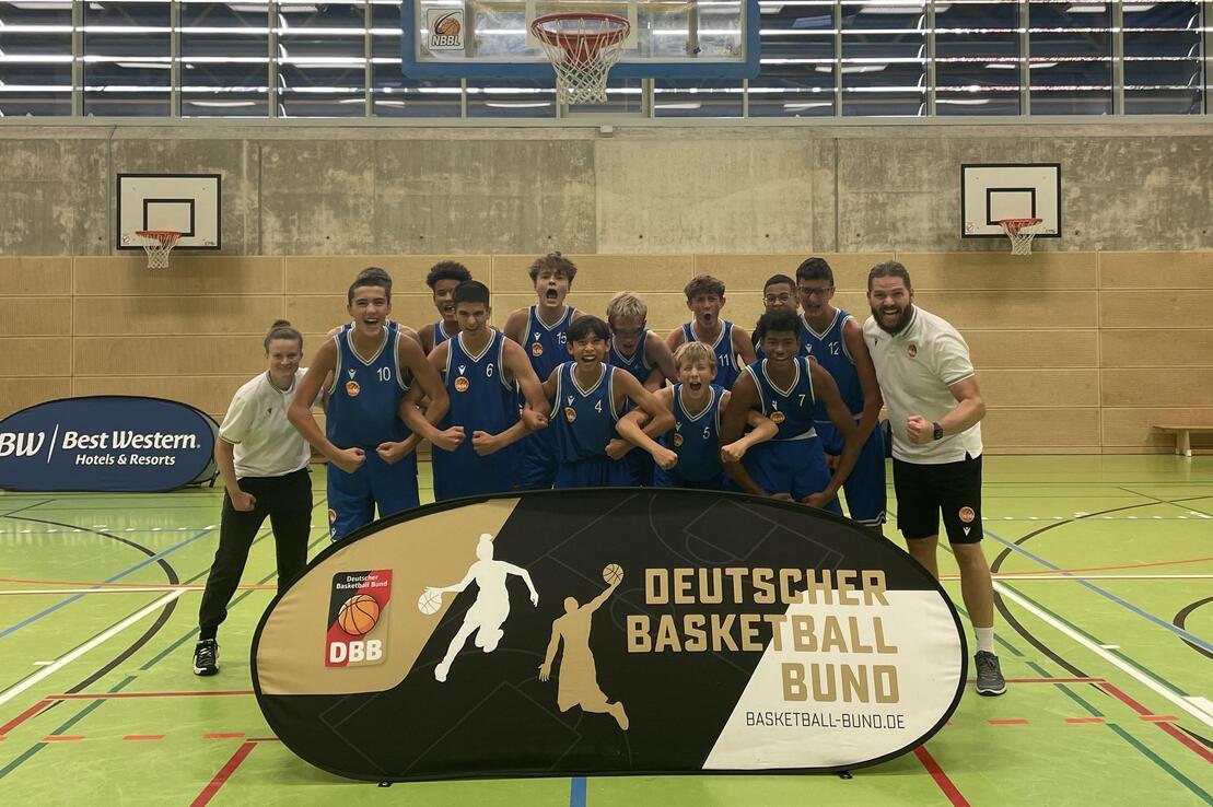 Gruppe von Basketballspielern in blauen Trikots posiert freudig vor einem Banner des Deutschen Basketball Bundes.