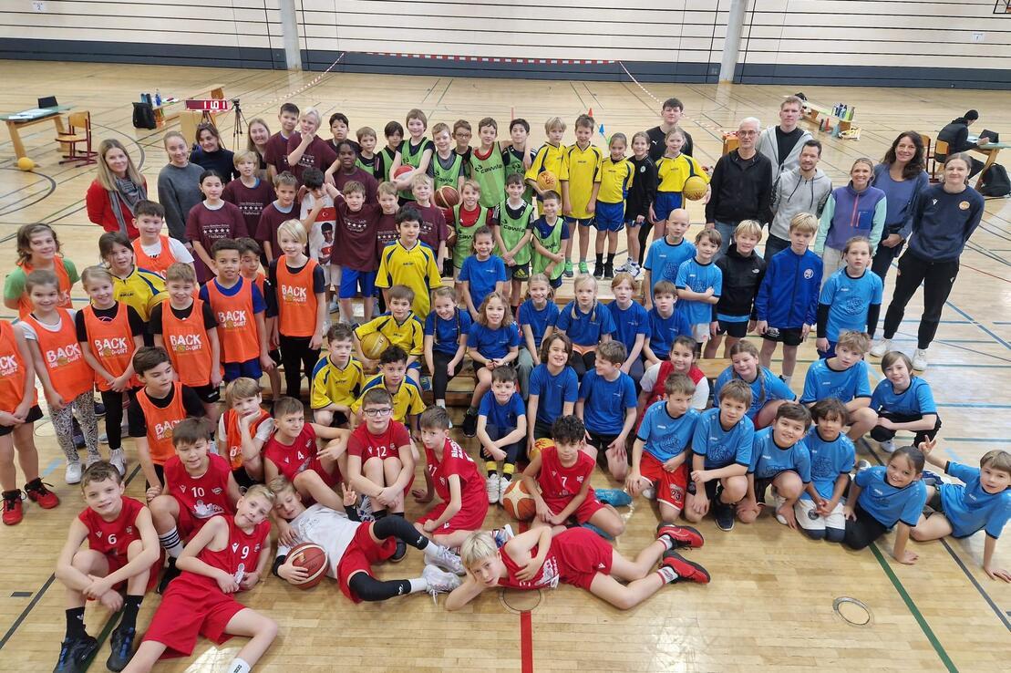 Gruppenshot von Kindern und Betreuern in Sportkleidung in einer Turnhalle, umgeben von Basketballb&auml;llen und Trainingsmaterial.