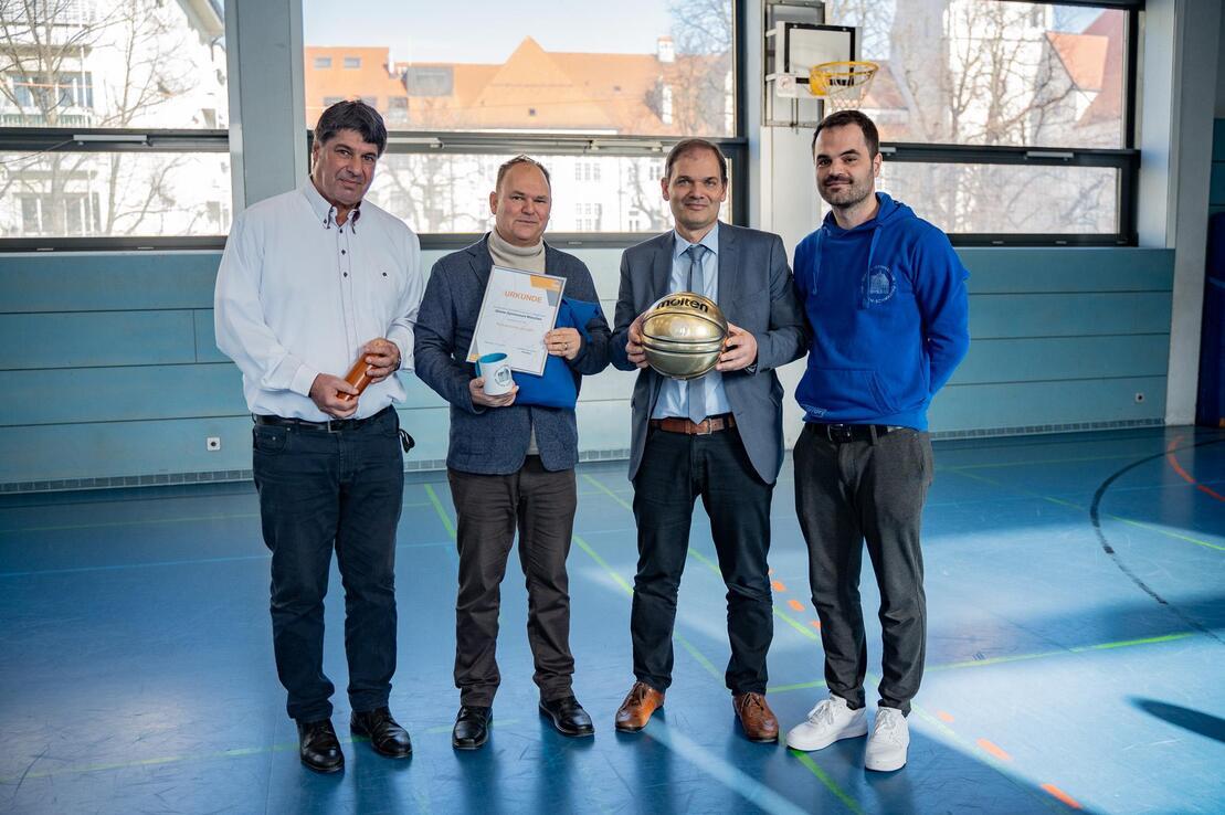 Vier Personen in Sportkleidung stehen in einer Turnhalle und halten Auszeichnungen und einen Basketball in der Hand.