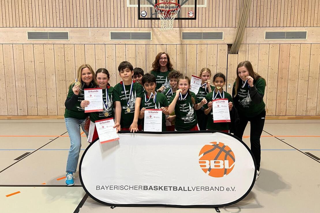 Gruppenfoto von Jugendlichen mit Medaillen und Urkunden vor einem Basketballkorb, gemeinsam mit einer Trainerin.