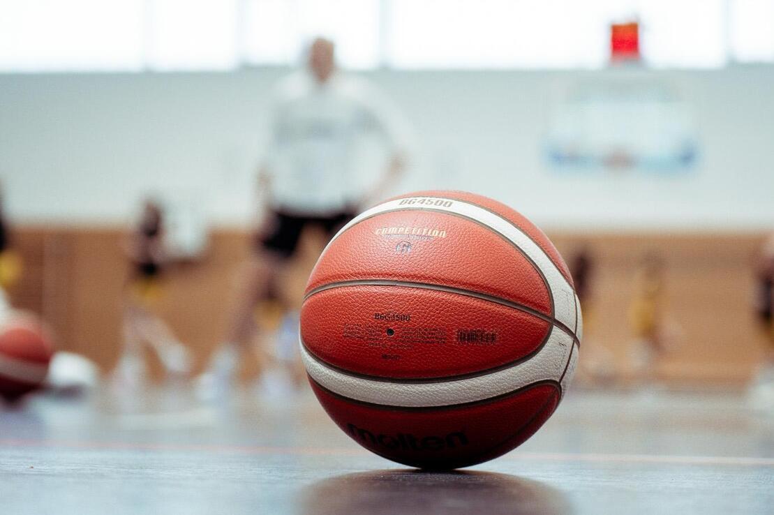Nahaufnahme eines Basketballs auf dem Hallenboden, Hintergrund unscharf mit Spielern und Basketballkorb.