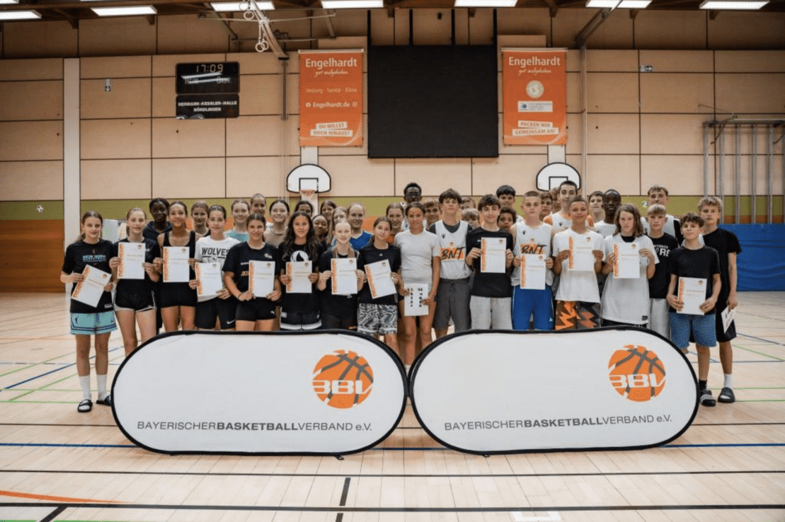 Gruppe von Jugendlichen mit Urkunden in einer Sporthalle, vor Bannern des Bayerischen Basketballverbands.