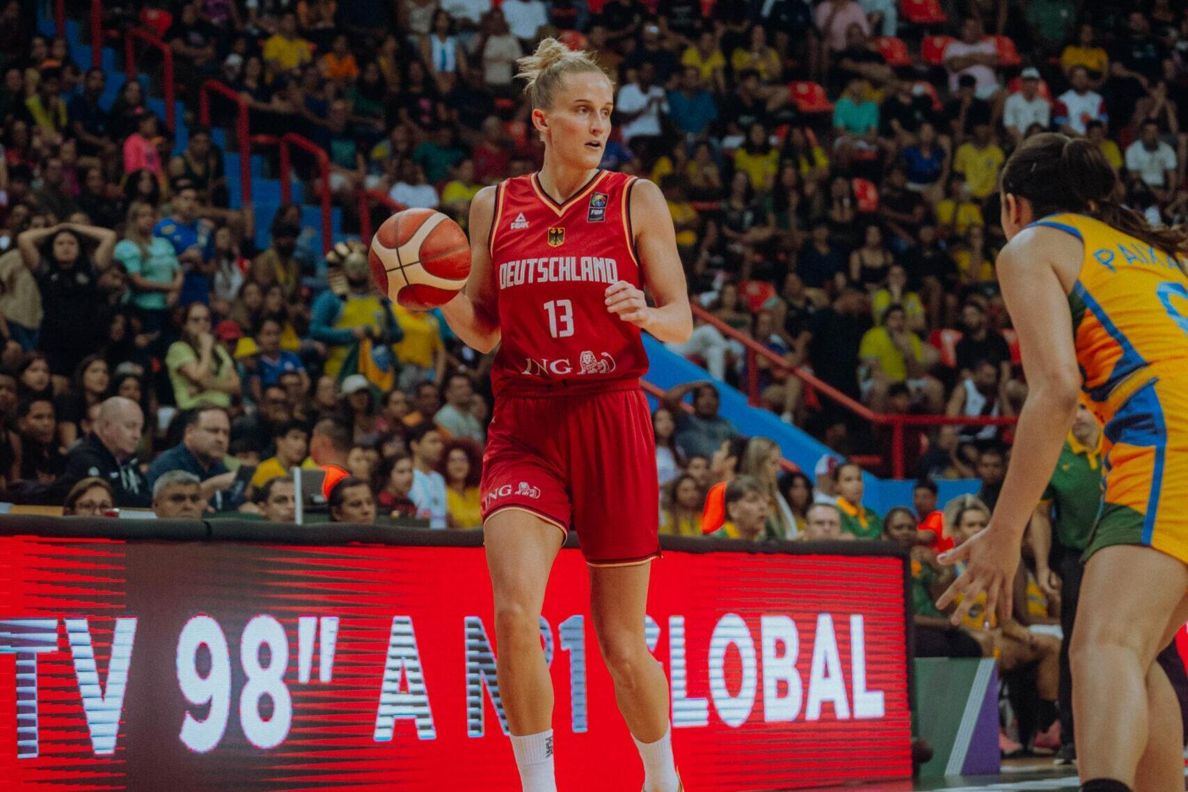Basketballspiel: Spielerin in rotem Trikot dribbelt den Ball auf dem Feld, Zuschauer im Hintergrund.