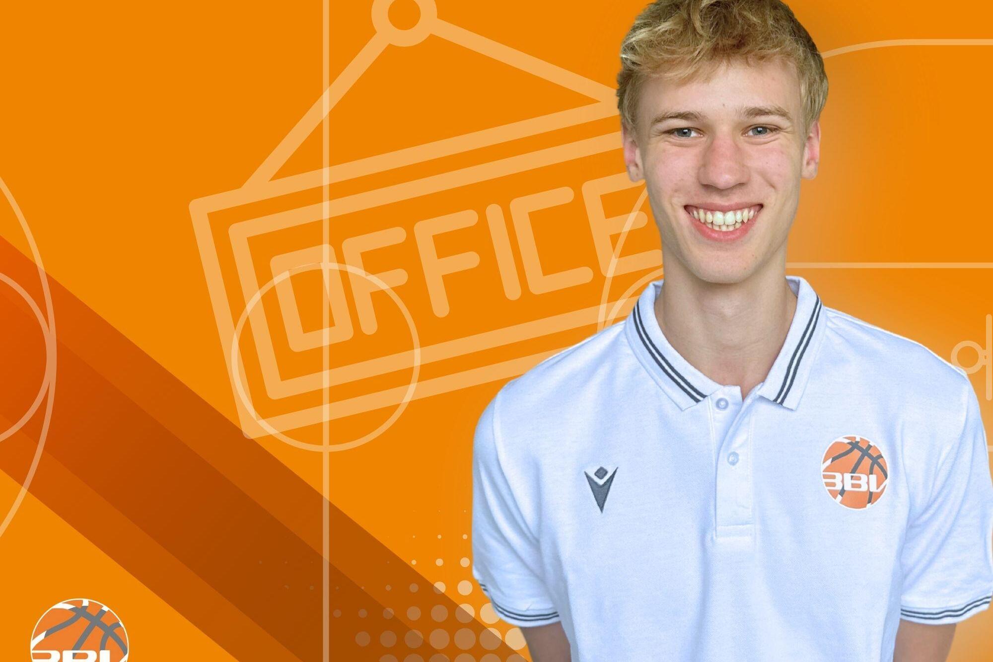 Junger Mann mit kurzem, blondem Haar, l&auml;chelt vor orangefarbenem Hintergrund mit &bdquo;OFFICE&ldquo;-Schild und Basketball-Logo.