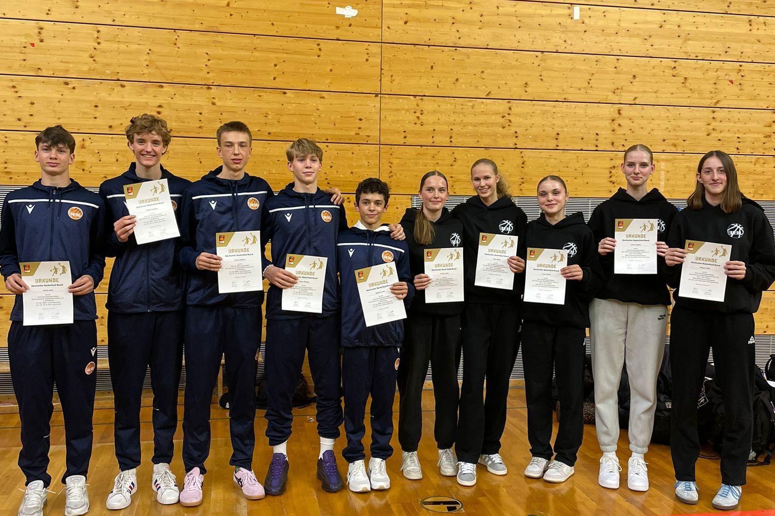 Gruppe von zehn Jugendlichen in Sportkleidung h&auml;lt Urkunden in einer Turnhalle mit Holzverkleidung.