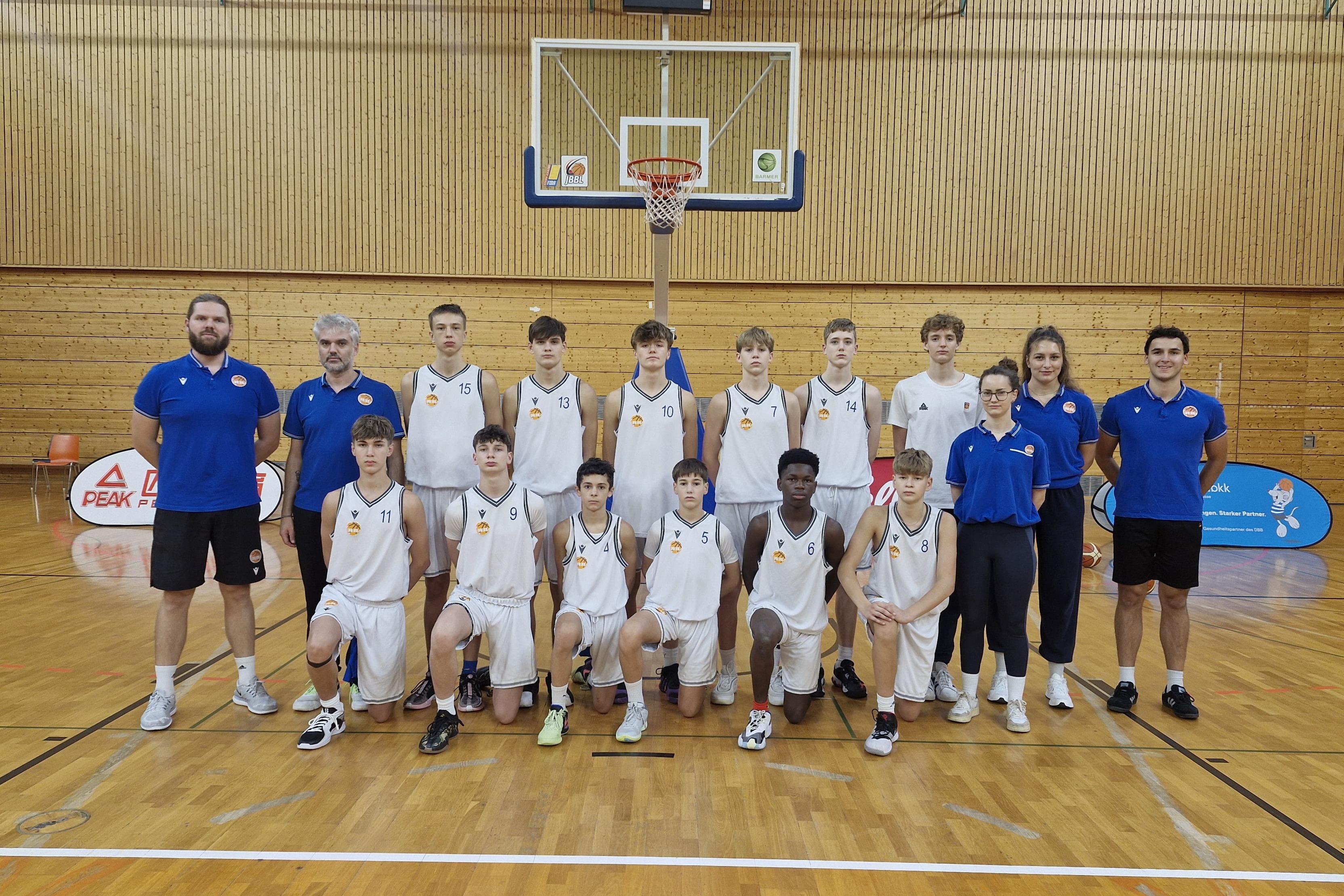 Mannschaftsfoto einer Jugend-Basketballmannschaft mit Trainern in Sportkleidung auf einem Basketballplatz.