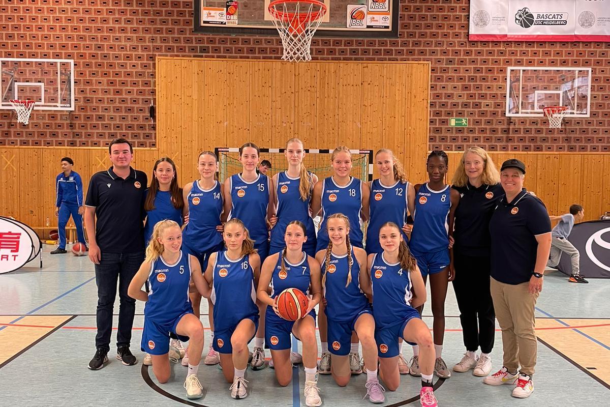 Foto einer Basketballmannschaft in blauen Trikots, stehend und kniend, mit Trainer in einer Sporthalle.