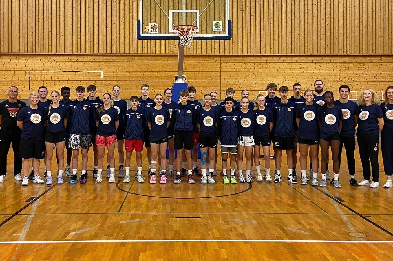 Gruppe von Basketballspielern und -trainern in Sportbekleidung, aufgestellt in einer Sporthalle vor einem Basketballkorb.