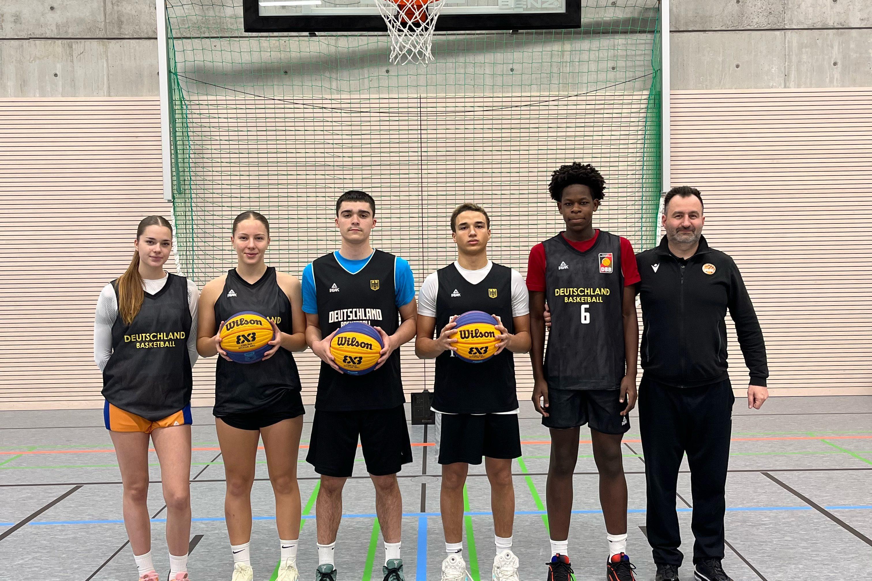 Sechs Basketballspieler in Sportkleidung stehen vor einem Basketballkorb in einer Halle und halten B&auml;lle in den H&auml;nden.