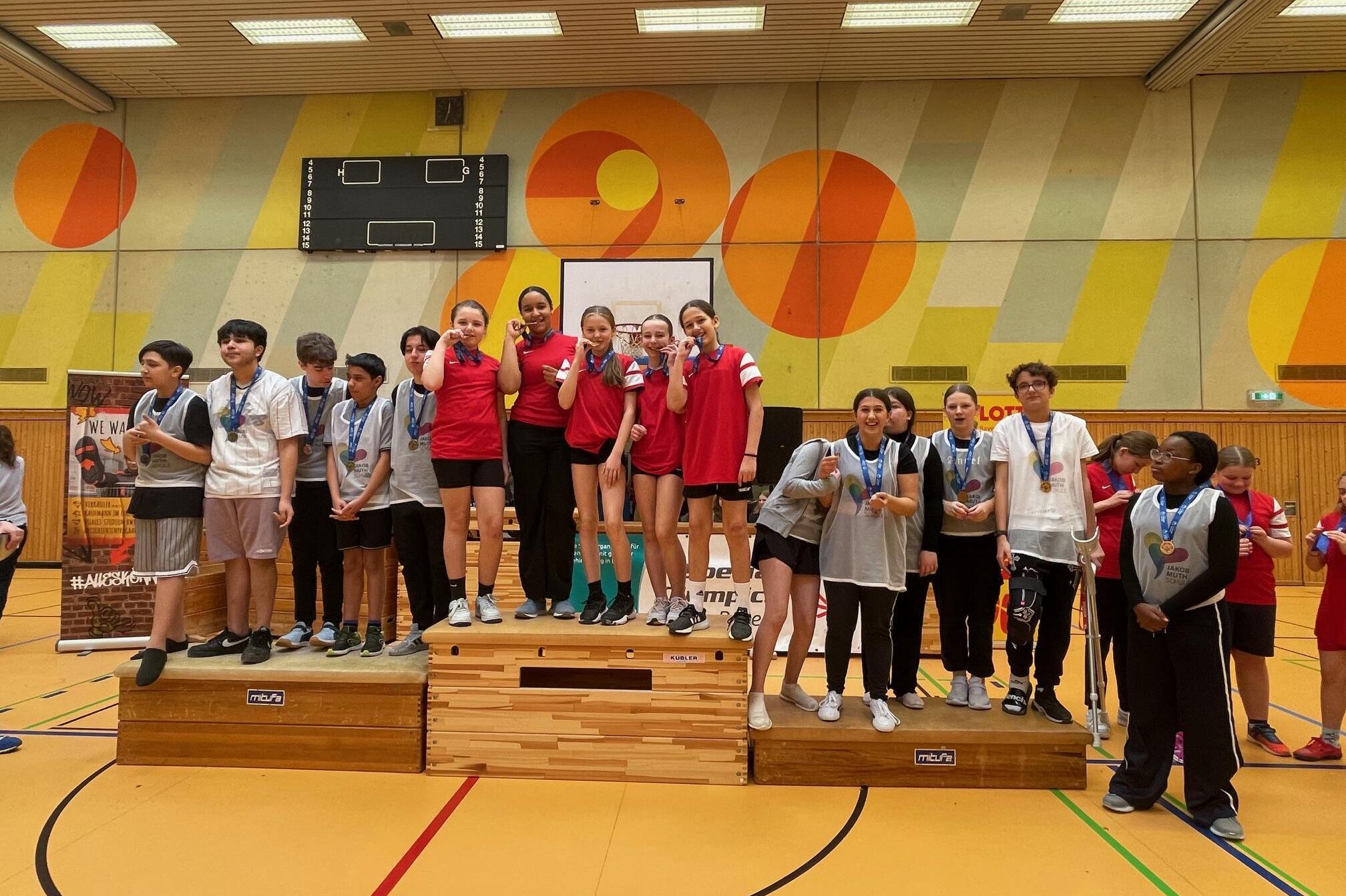 Gruppe von Jugendlichen auf dem Siegertreppchen mit Medaillen in einer Sporthalle. Farbig gestaltete Wand im Hintergrund.