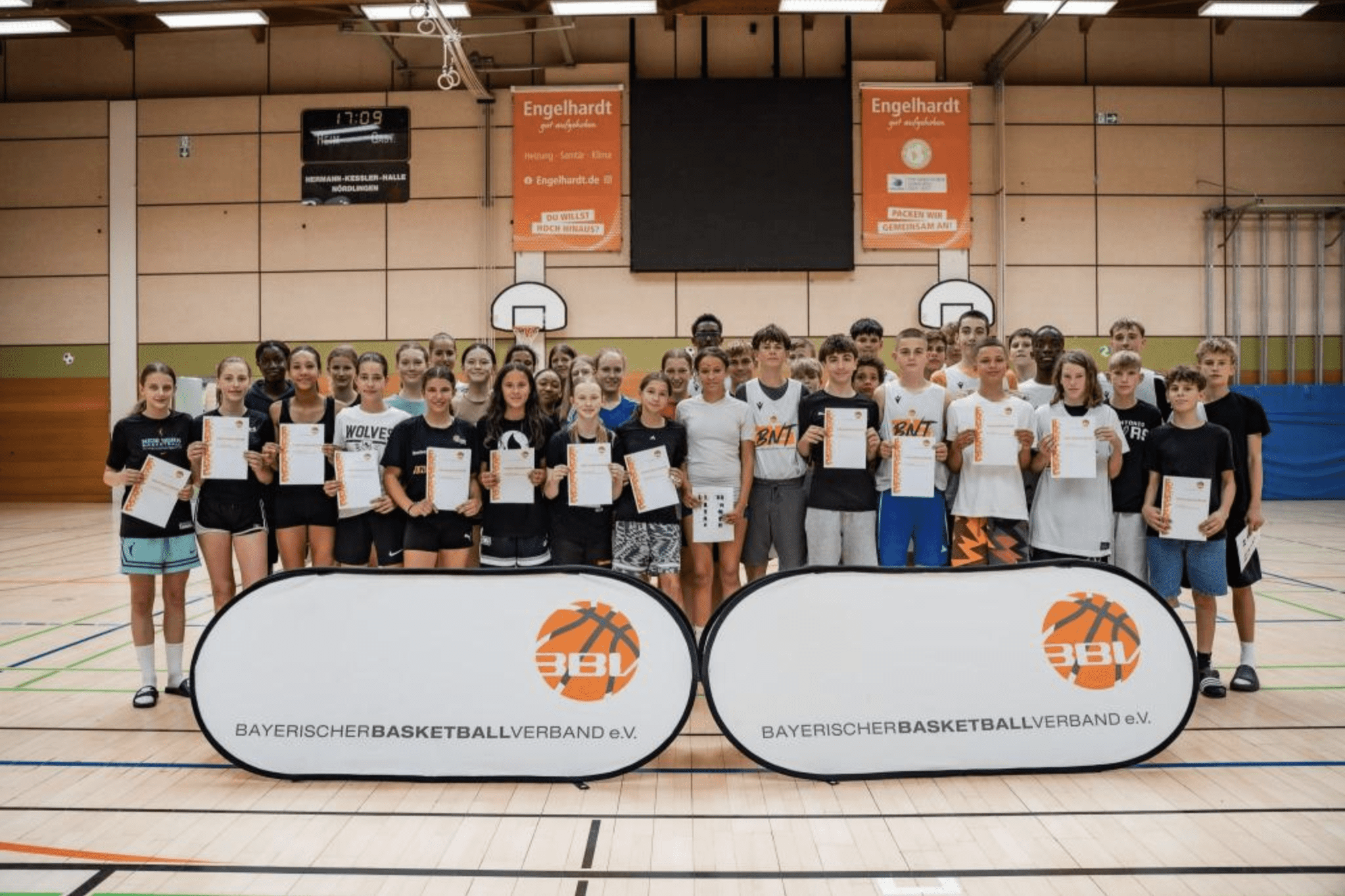 Gruppe von Jugendlichen mit Urkunden in einer Sporthalle, vor Bannern des Bayerischen Basketballverbands.