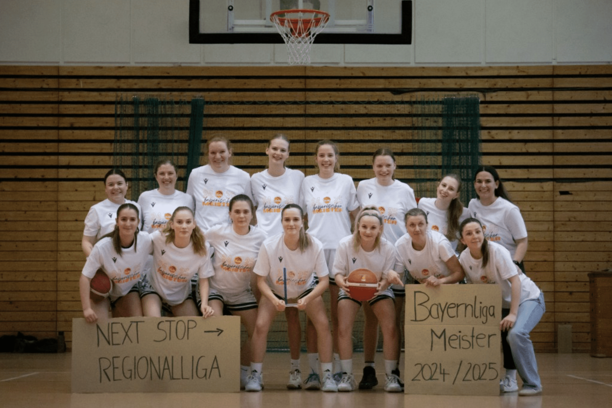 Frauen-Basketballmannschaft posiert in T-Shirts vor einem Basketballkorb, mit Schildern zu Regional- und Bayernliga-Meisterschaft.