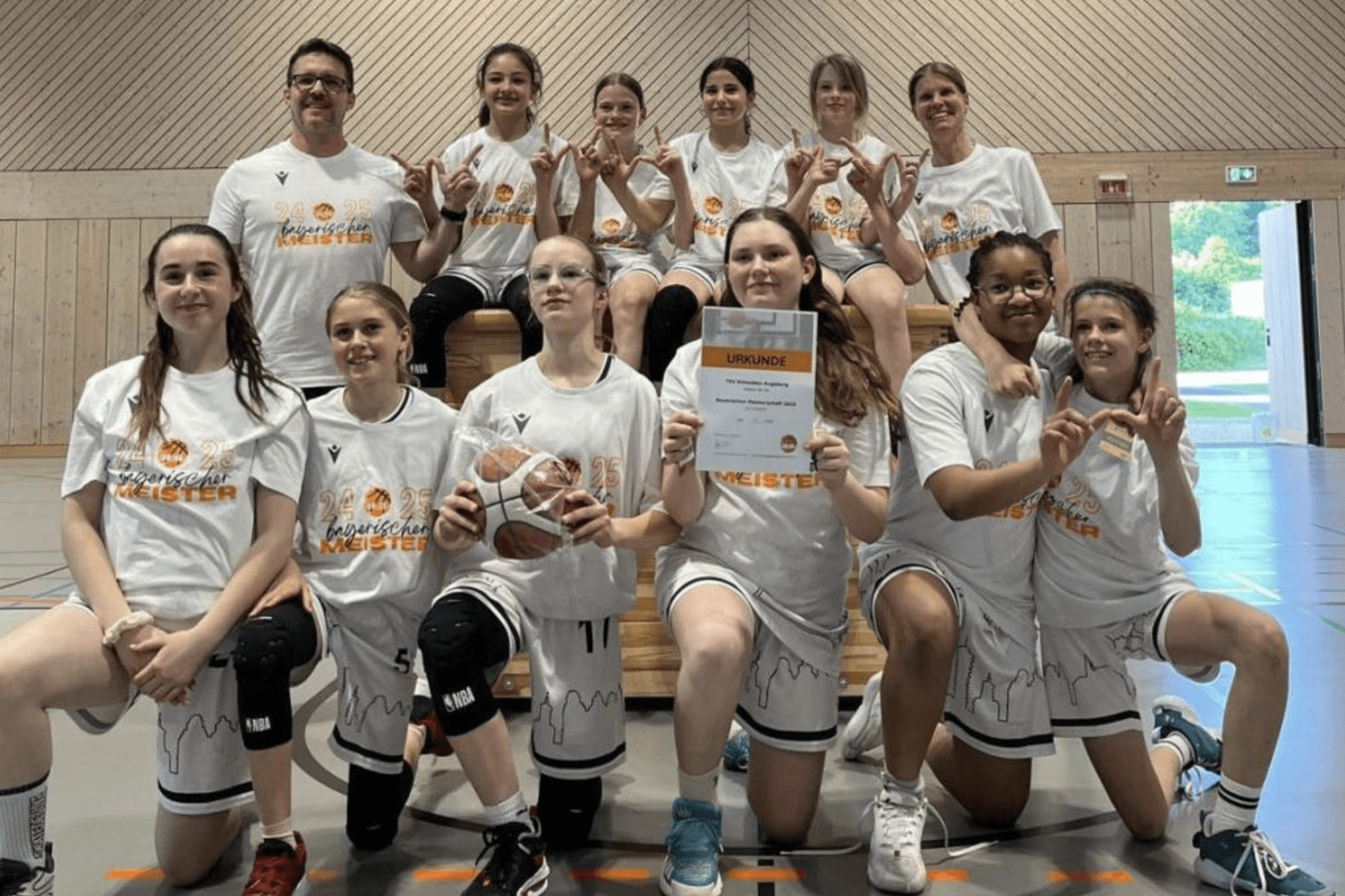 Gruppenshot eines Basketballteams in wei&szlig;en T-Shirts, stolz mit Urkunde und Basketball auf einem Holzblock.