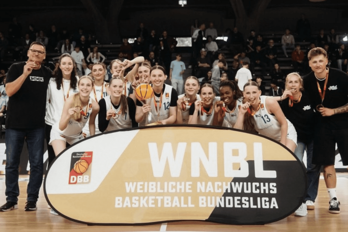 Feiernde Basketballmannschaft mit Medaillen und Pokal vor einem Banner der WNBL in einer Halle.