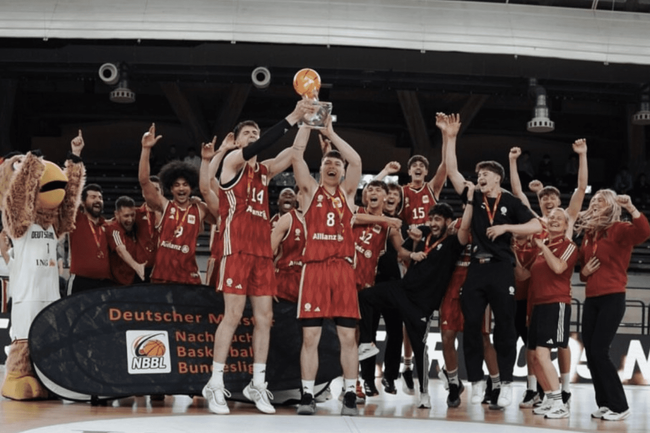 Basketballmannschaft feiert mit Pokal, Menschen jubeln, in Sportbekleidung auf dem Platz, Maskottchen anwesend.