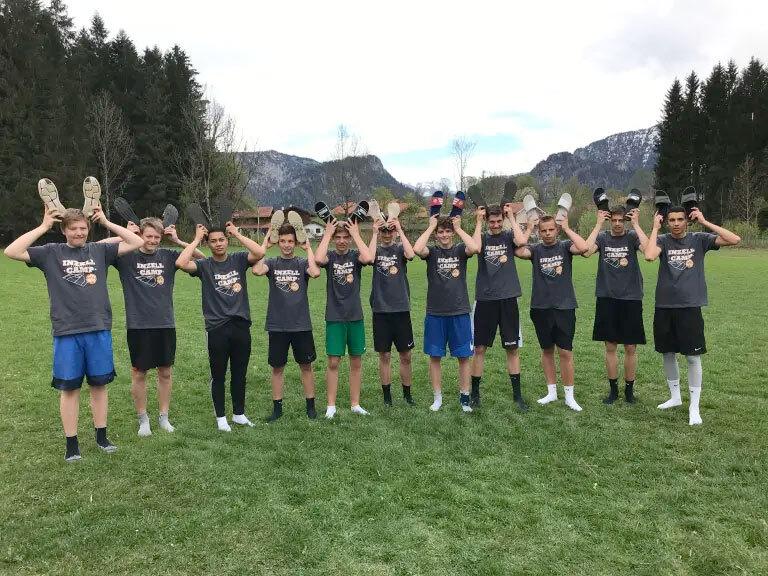 Gruppe von 13 Jugendlichen in Sportkleidung, die Schuhe &uuml;ber den K&ouml;pfen auf einem Rasenfeld halten.