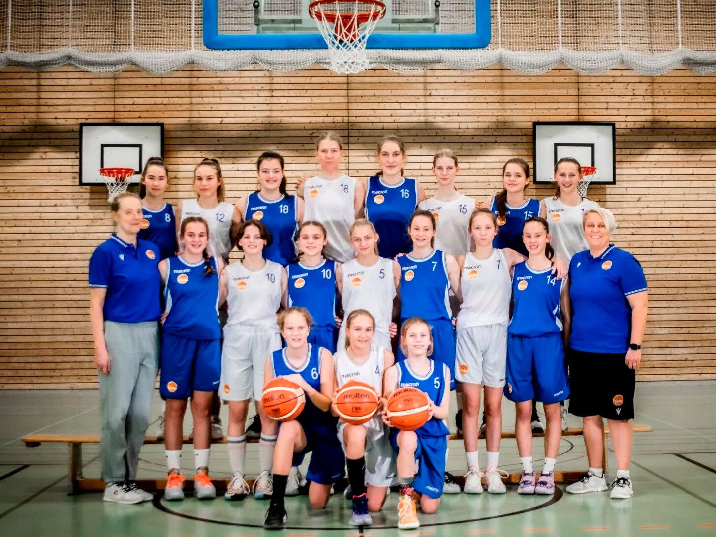 Gruppenfoto eines Jugend-Basketballteams mit Trainerinnen in Sportkleidung, auf einem Basketballfeld in einer Halle.