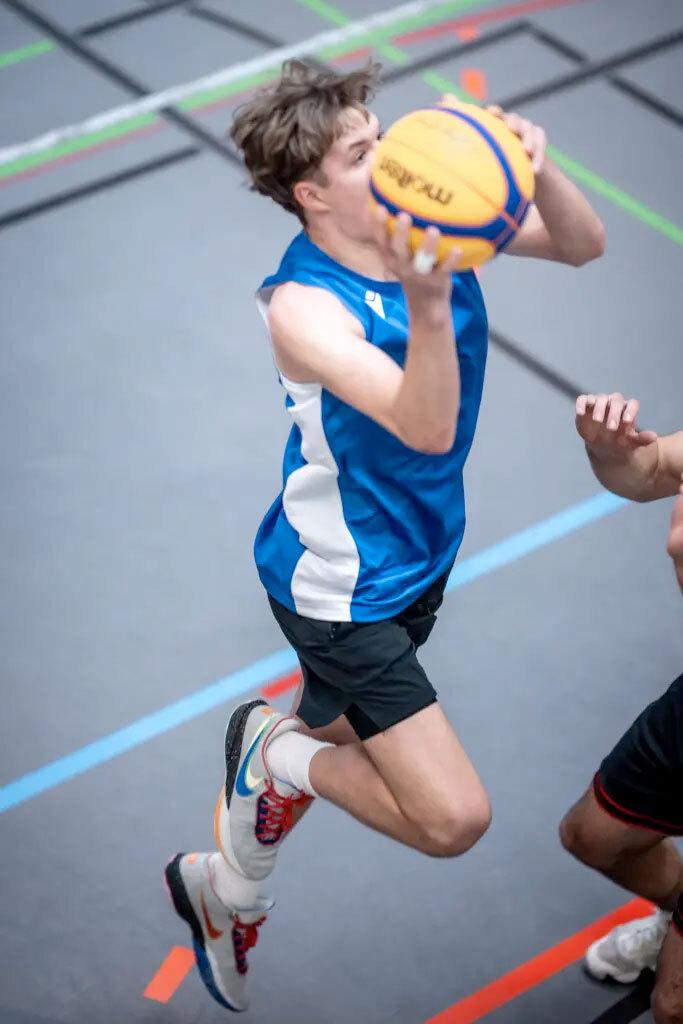 Spieler in blauem Trikot springt zum Wurf mit einem Basketball in einer Sporthalle. Ein weiterer Spieler ist in der N&auml;he.