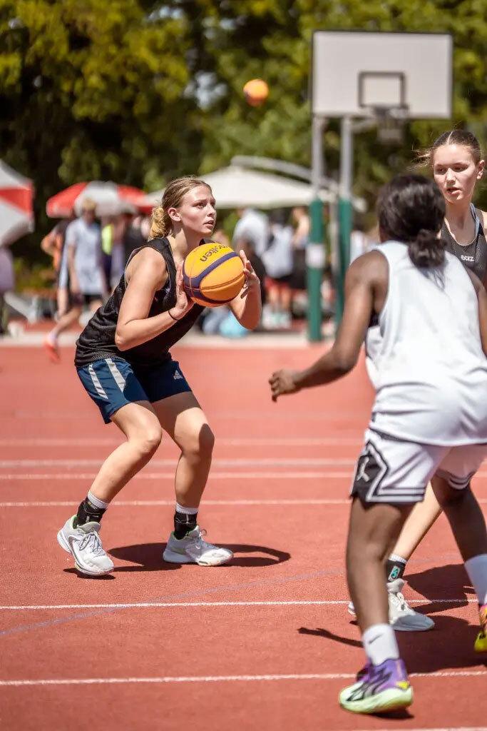 Ein M&auml;dchen mit Ball in der Hand bereitet sich auf einen Wurf auf einem Basketballplatz vor, weitere Spieler sind im Hintergrund.