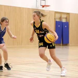 Basketballspiel: Spielerin in schwarzer Uniform dribbelt mit Ball, w&auml;hrend eine Gegenspielerin sie verfolgt.