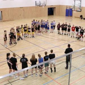 Gruppierung junger weiblicher Basketballspieler im Sporthallen, w&auml;hrend ein Trainer an sie herantritt.