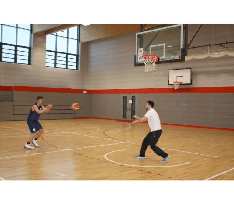 Zwei M&auml;nner spielen Basketball in einer Sporthalle; einer wirft den Ball, w&auml;hrend der andere ihn f&auml;ngt.