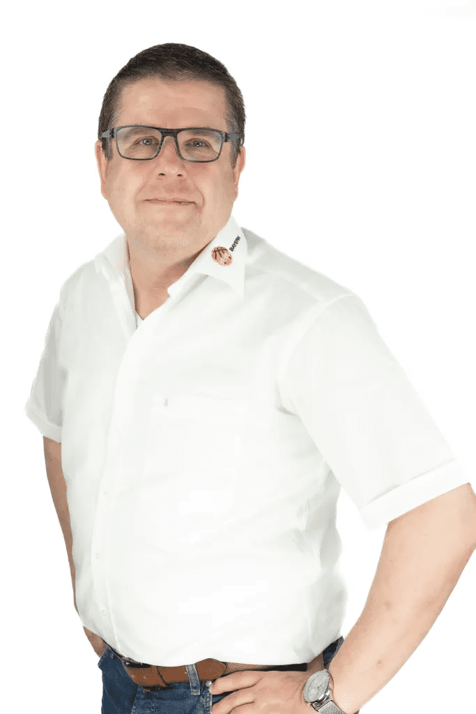 Mann mit Brille in wei&szlig;em Hemd l&auml;chelt, steht mit verschr&auml;nkten H&auml;nden vor wei&szlig;em Hintergrund.